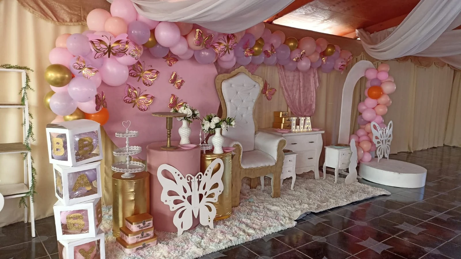 Montaje de celebración con globos rosados y dorados, mariposas, flores y una silla blanca decorativa.