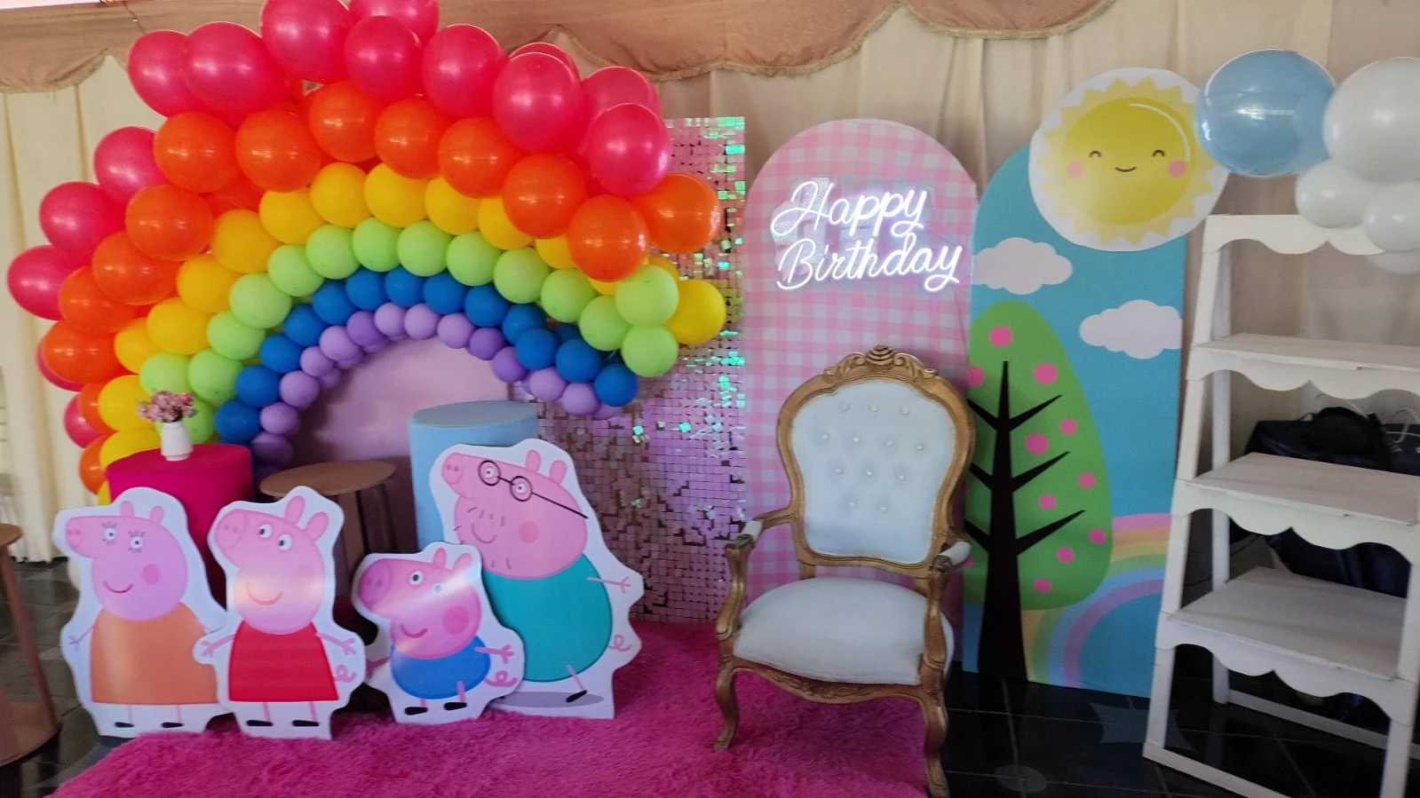 Decoración de cumpleaños con arcoíris de globos, figuras de Peppa Pig, silla blanca con marco dorado y letrero Happy Birthday.