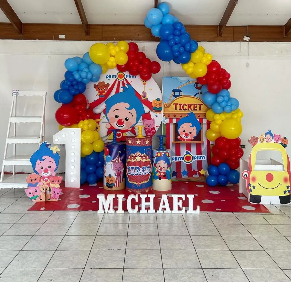 Montaje de cumpleaños infantil con temática de circo, globos rojos, amarillos y azules, figuras decorativas y un número uno luminoso.