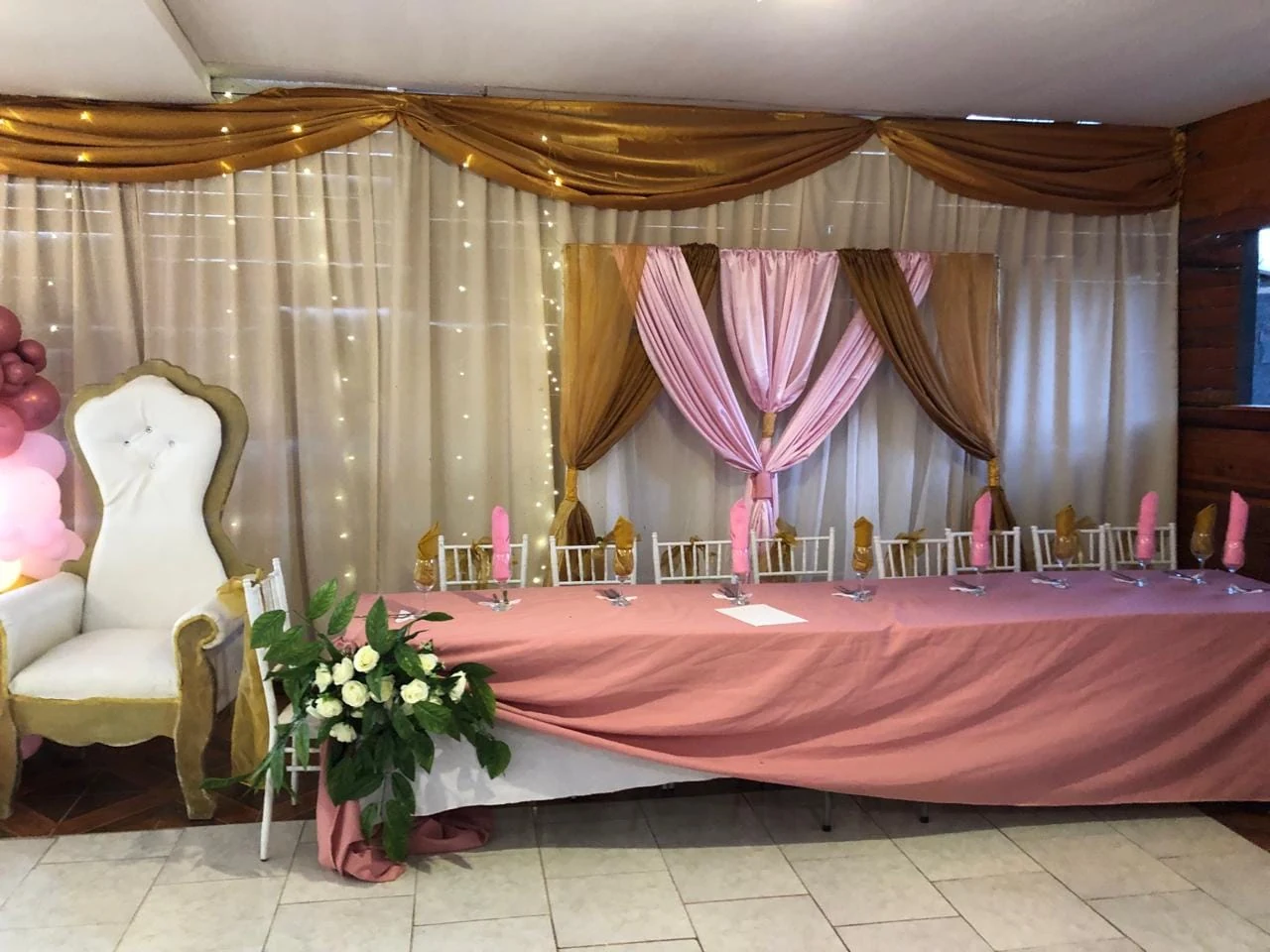 Mesa de cumpleaños con mantelería rosa, detalles dorados, sillas blancas y telas decorativas rosas y doradas.