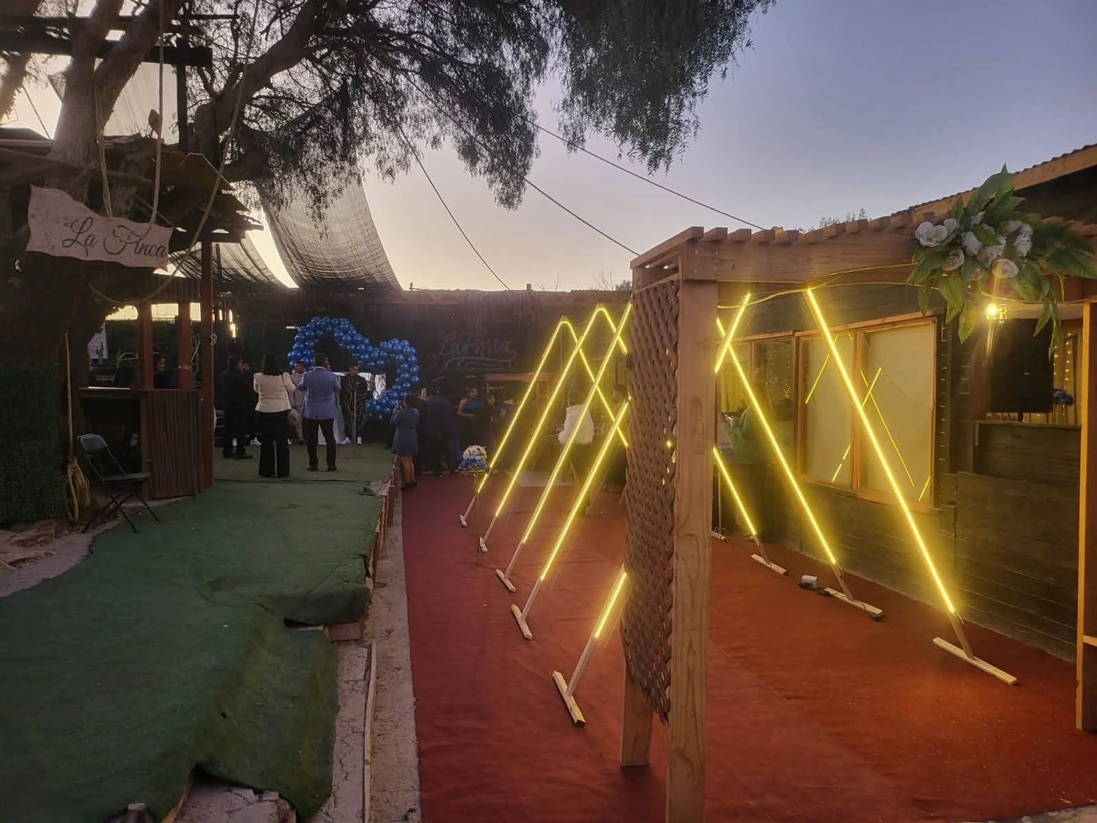 Exterior de Manantial durante una cena corporativa, con invitados al fondo, construcciones de madera y luces amarillas al atardecer.