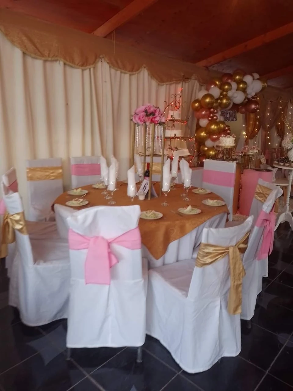 Mesa redonda con sillas blancas, lazos rosados, copas y un centro floral en un salón decorado.