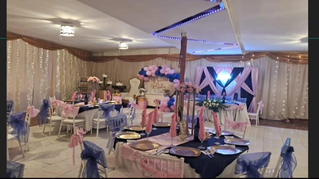 Salón preparado con mesas redondas, telas claras, detalles rosados y violetas e iluminación en tonos azules.
